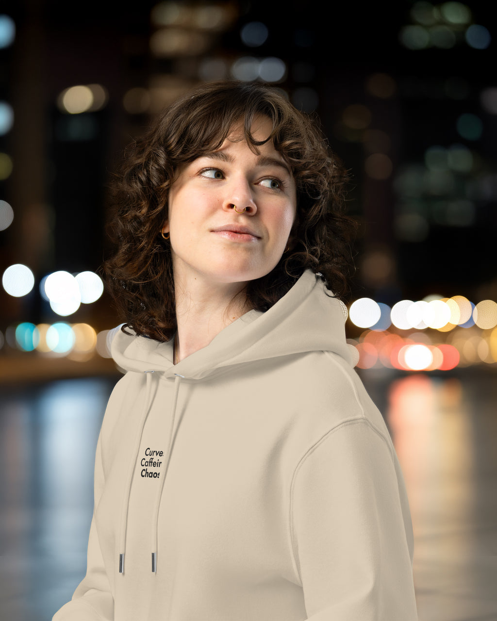 Person wearing a beige hoodie with text, standing against a blurred cityscape background at night.