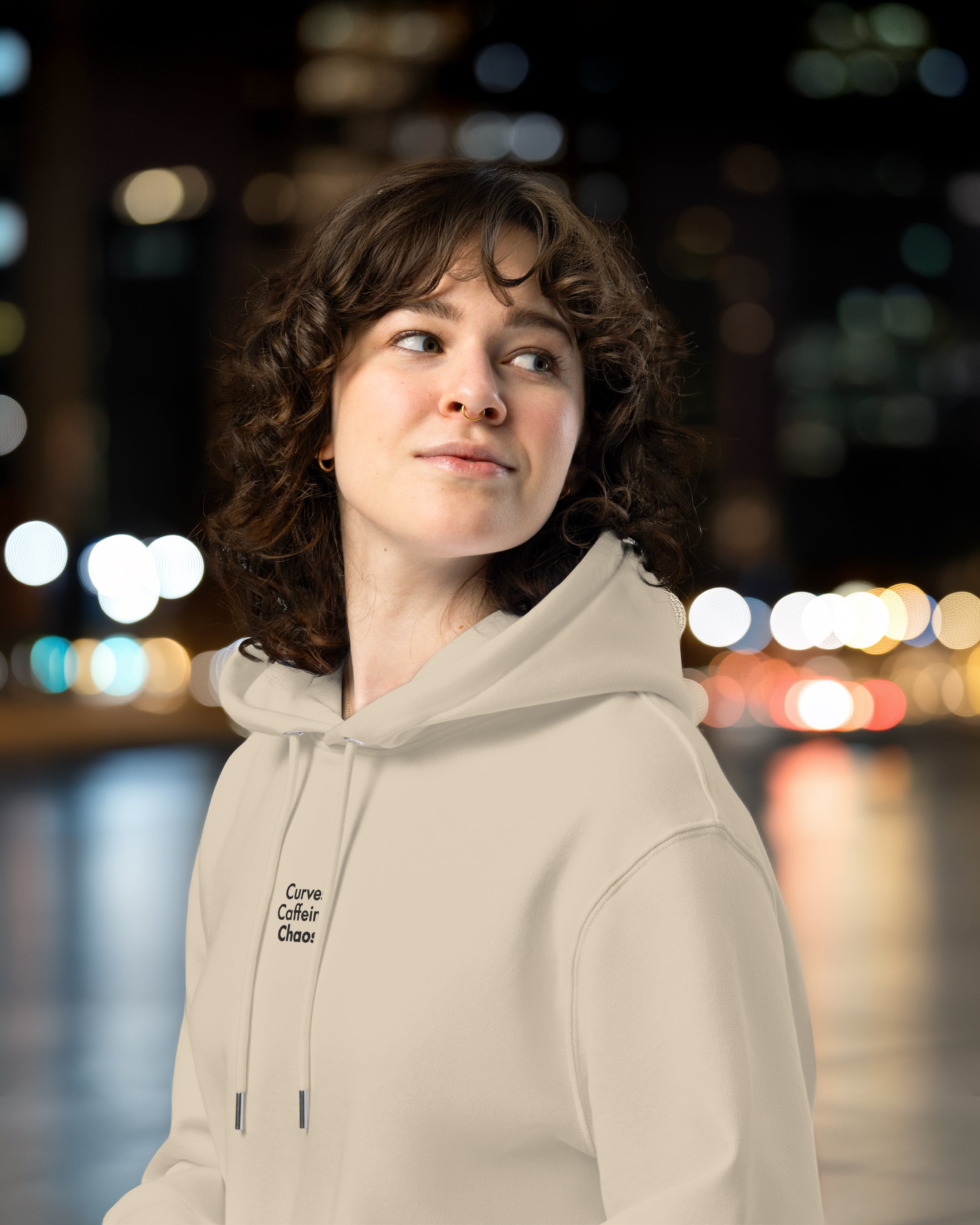 Person wearing a beige hoodie with text, standing against a blurred cityscape background at night.