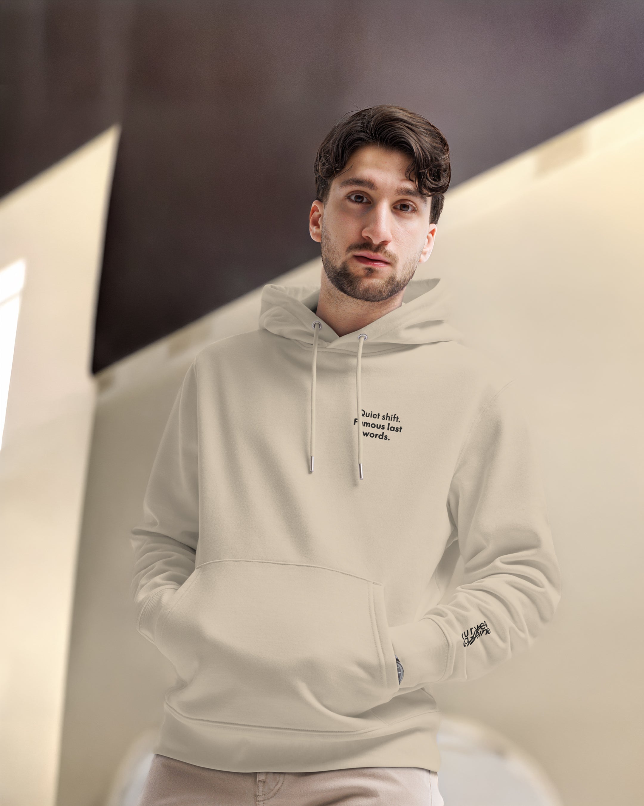 Man wearing a beige hoodie with text, standing indoors against a neutral background