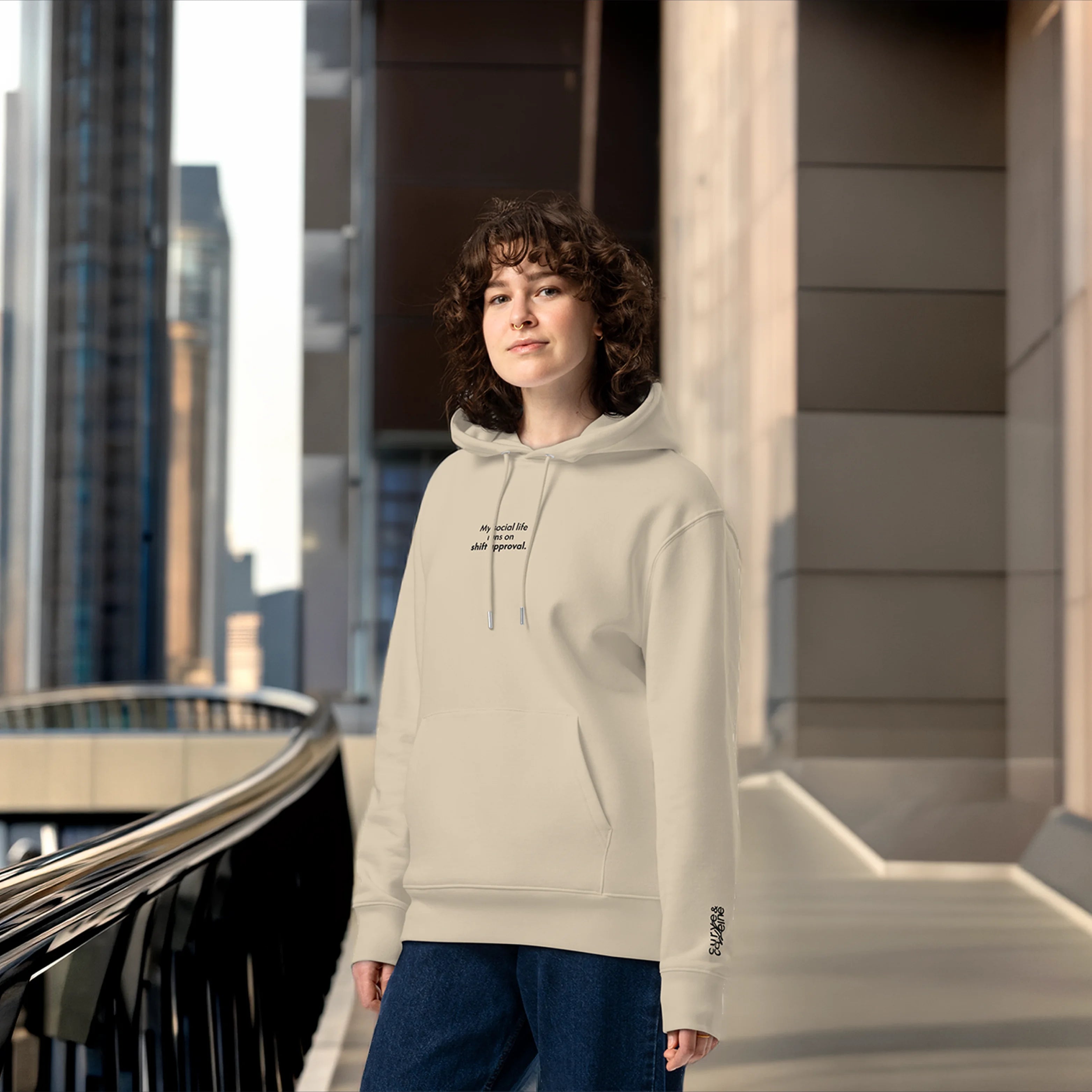 Person wearing a beige hoodie with text, standing outdoors in an urban setting.